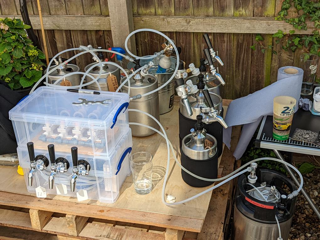 A collection of miniature kegs atop a wooden pallet. A translucent plastic box sits atop the pallet with three beer taps attached to its front. Another plastic box sits atop the first box.
